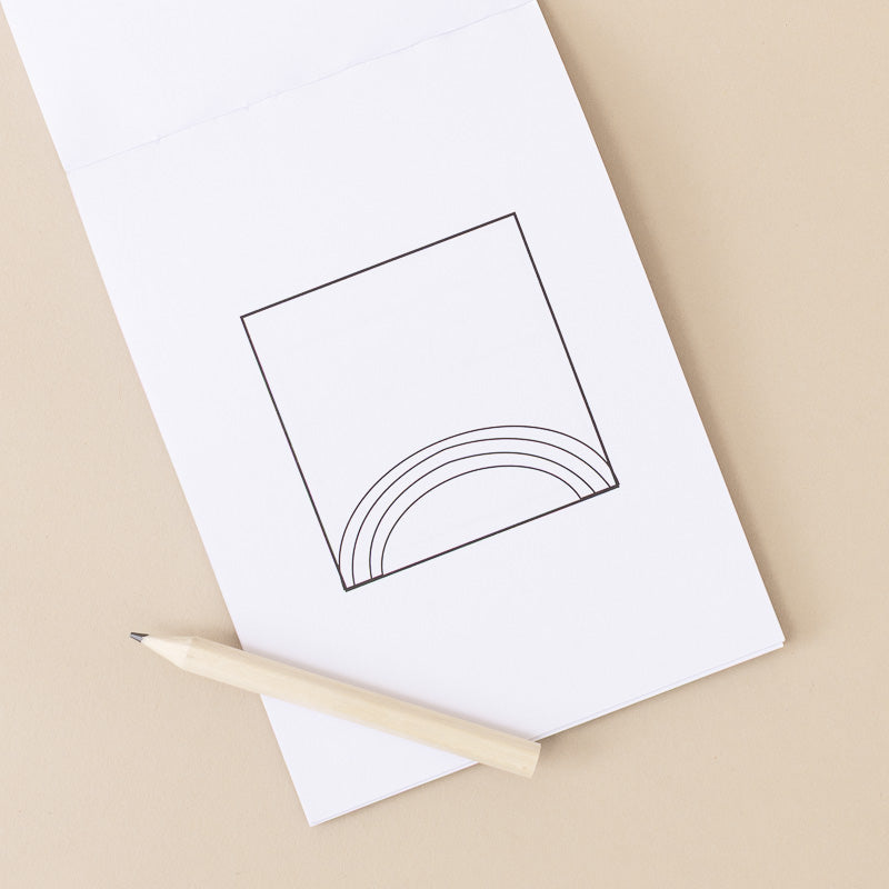 A white sketchbook or notepad with a simple black line drawing of a square and rainbow, placed on a beige surface with a light wooden pencil resting across the bottom.