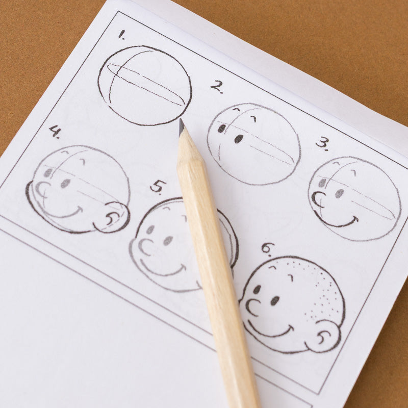 A close-up of a printed face-drawing instruction sheet with numbered steps showing how to sketch a round head, with a wooden pencil resting diagonally across it on a brown surface.