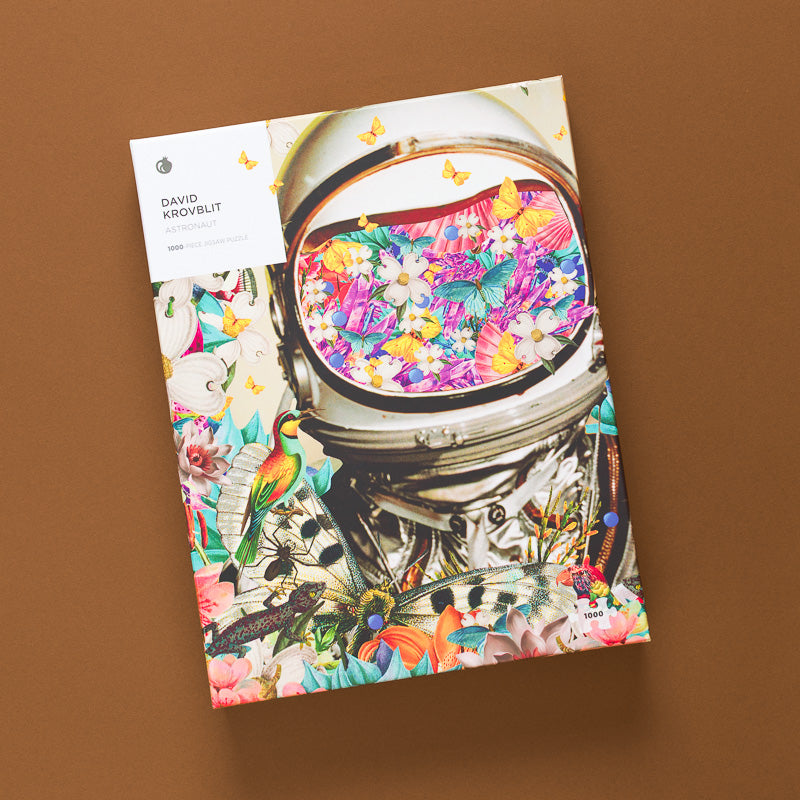 The puzzle 'David Arovbit: The Universe' by David Arovbit is displayed upright on a solid brown background. The cover features a highly detailed, colorful illustration of an astronaut's helmet filled with flowers, surrounded by butterflies, birds, and other nature motifs.