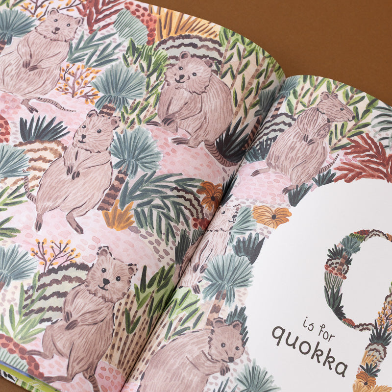An open children's book showing a whimsical illustration of quokka animals among tropical plants on a pink background, with a white page partially visible on the right and the word 'quokka' printed at the bottom.