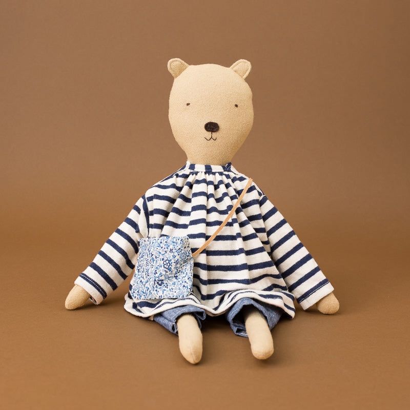A plush teddy bear doll with a beige fabric face and limbs, wearing a navy and white striped long-sleeve top and denim-style pants, with a small blue-and-white patterned crossbody bag, posed against a solid brown background.