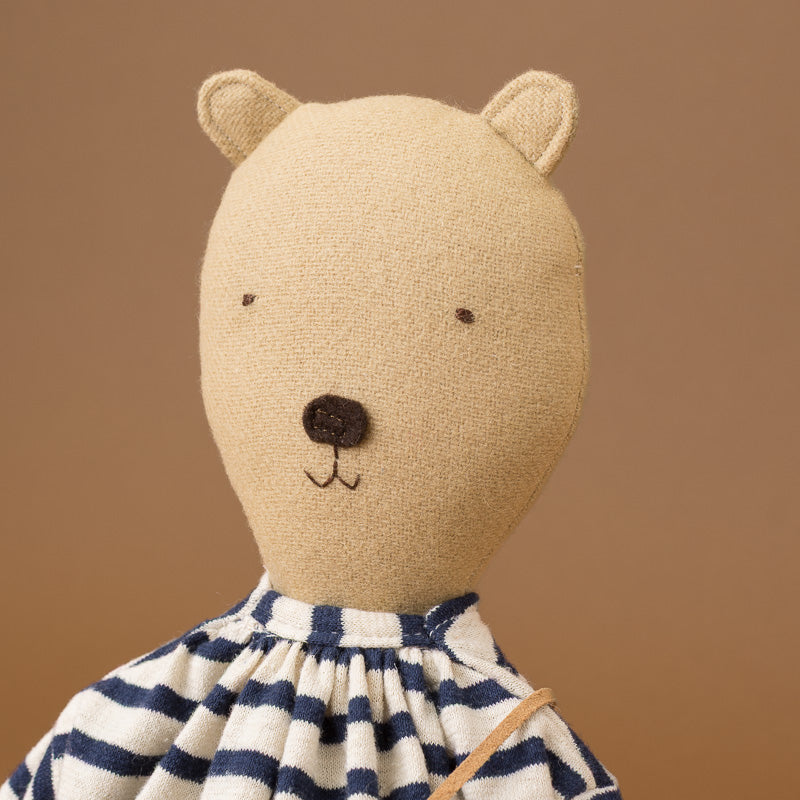 A close-up of a teddy bear doll with a beige linen-textured face and ears, a dark brown embroidered nose and mouth, and a navy-and-white striped dress against a warm brown background.