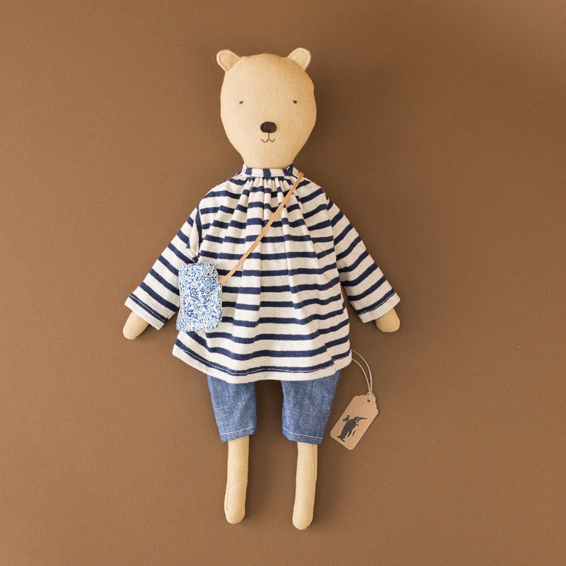 A plush teddy bear doll wearing a navy and white striped long-sleeve shirt, denim shorts, and a small blue patterned crossbody bag, posed against a solid brown background.