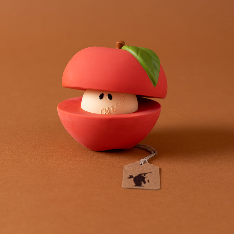 A novelty lip balm container shaped like a red apple with a green leaf, shown open to reveal a cream-colored balm inside. A small paper hangtag with a black logo is attached.