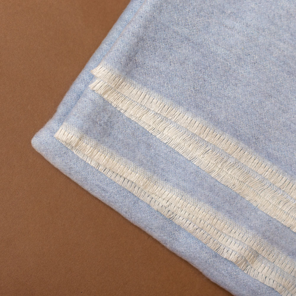 aspen-throw-hydrangea Light blue fabric with white fringe trim on a brown background