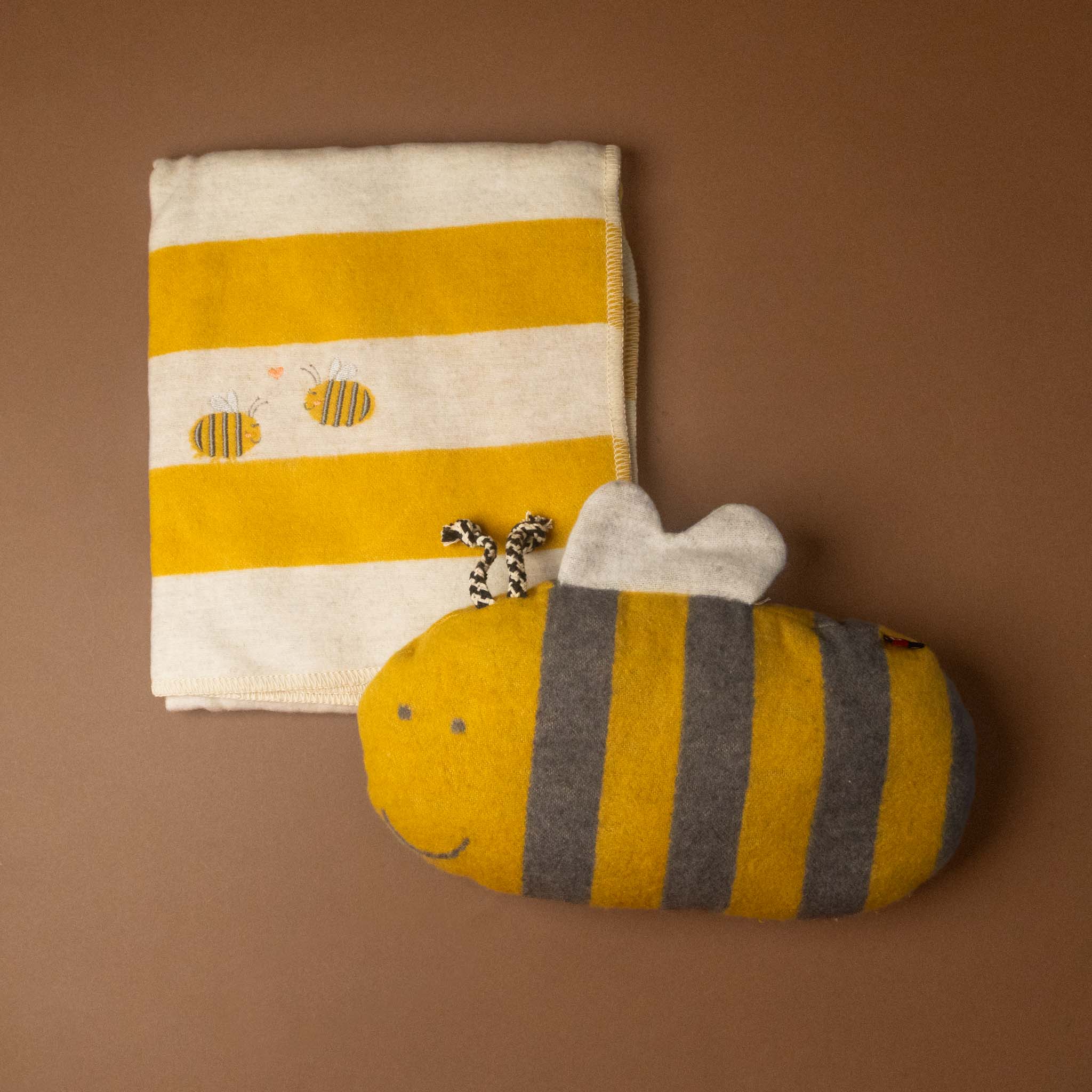 Striped yellow and white blanket with a bee design, accompanied by a bumble-bee-cushion-pillow on a brown background.