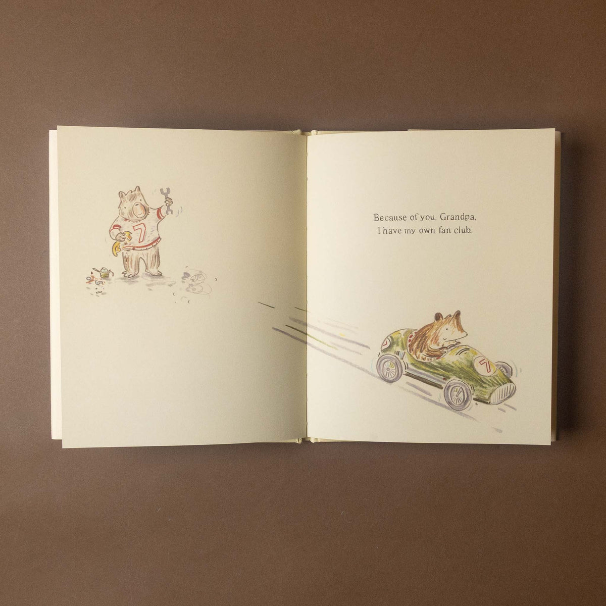 An open children's book laid flat on a brown surface, showing cream-colored pages with whimsical illustrations of a dog in a green race car and a cat holding a flower. The right page includes the text, 'Because of you, Grandpa, I have my own bus club.