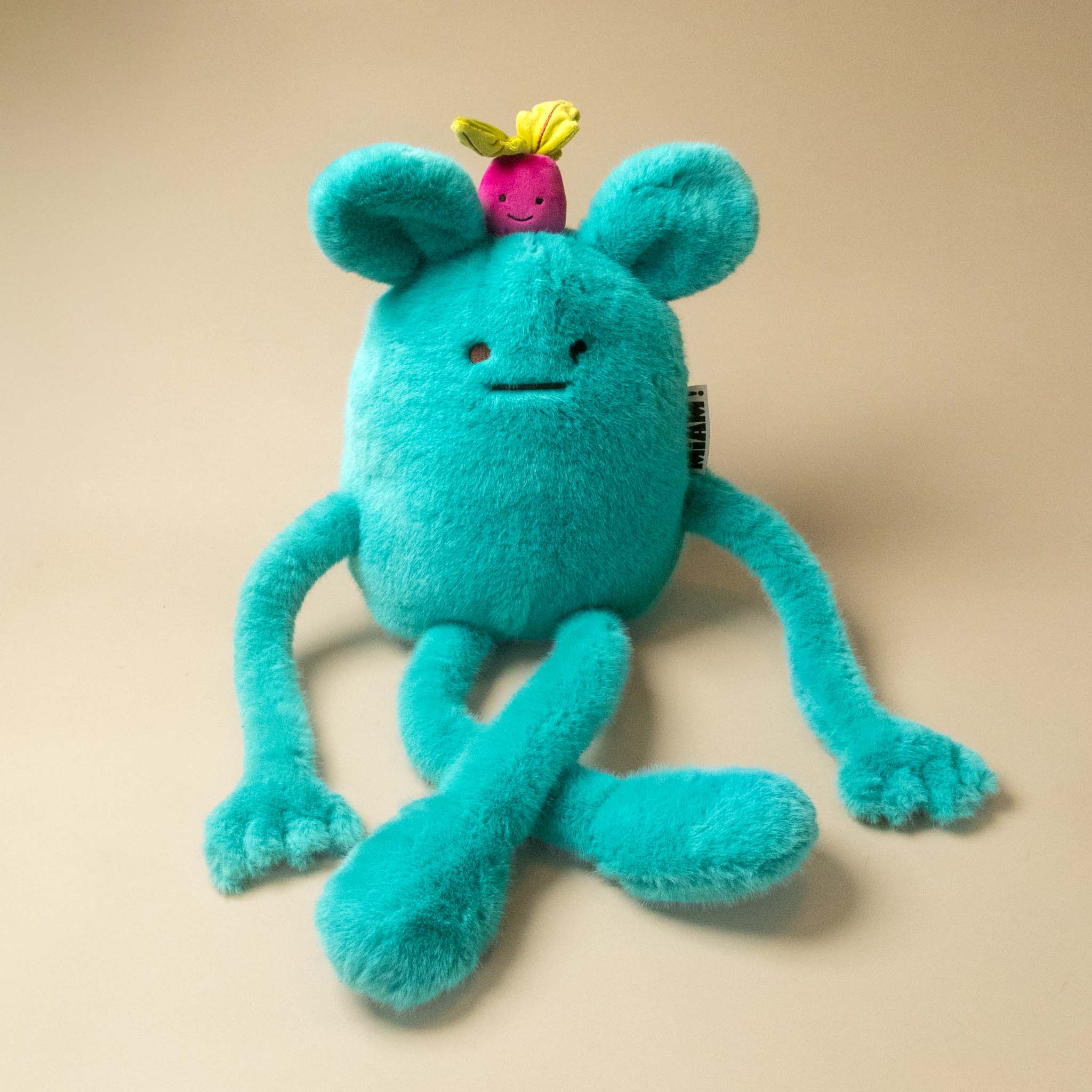 A turquoise blue plush billie-radish-grand toy with long limbs, a small pink flower on its head, and a hangtag, photographed on a plain beige background.