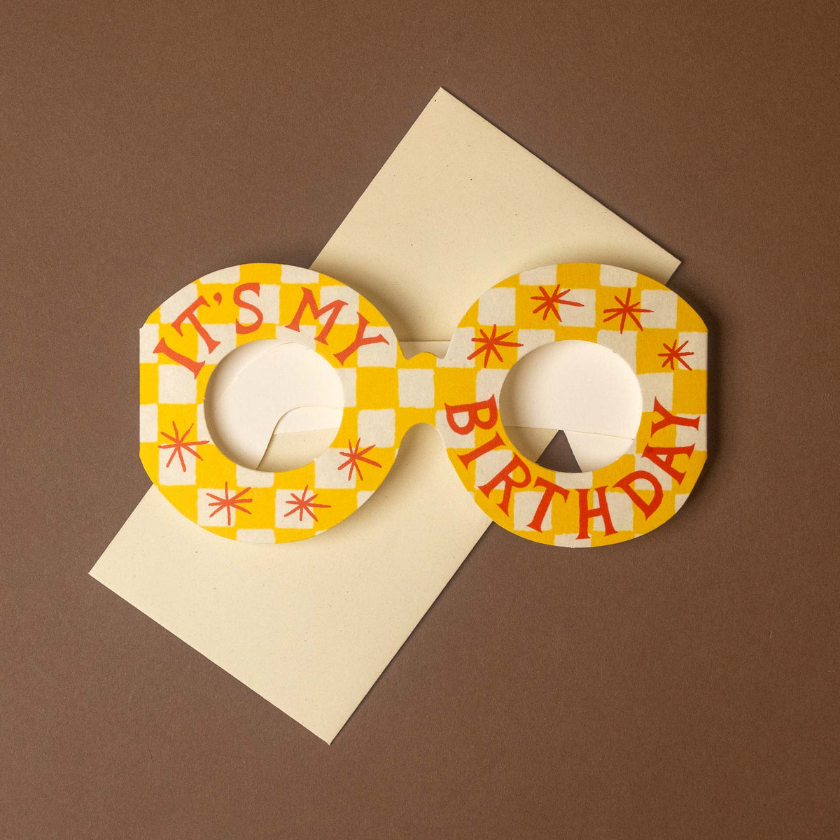 A pair of novelty round sunglasses with a yellow and white checkerboard pattern, red starburst accents, and the words 'IT'S MY BIRTHDAY' printed in red across the front. The sunglasses are displayed on top of a cream-colored envelope against a solid brown background.