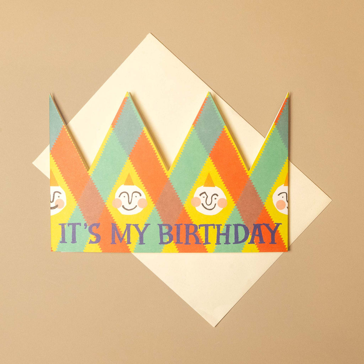 A colorful birthday card with a crown-shaped design and the text 'IT'S MY BIRTHDAY' in blue, layered over a cream envelope on a beige background.