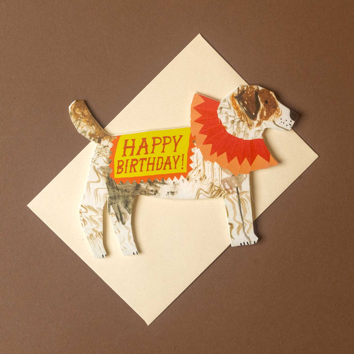 A handmade dog-shaped birthday card with a patterned white and brown body, a red collar, and a yellow belly patch that reads 'HAPPY BIRTHDAY!' in red letters, placed on a cream envelope over a brown background.