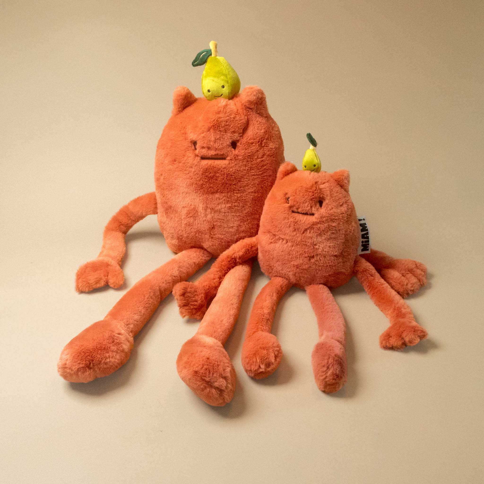 An orange plush britney-pear-petit and grand toy with a small yellow pear on its head, photographed on a plain beige background