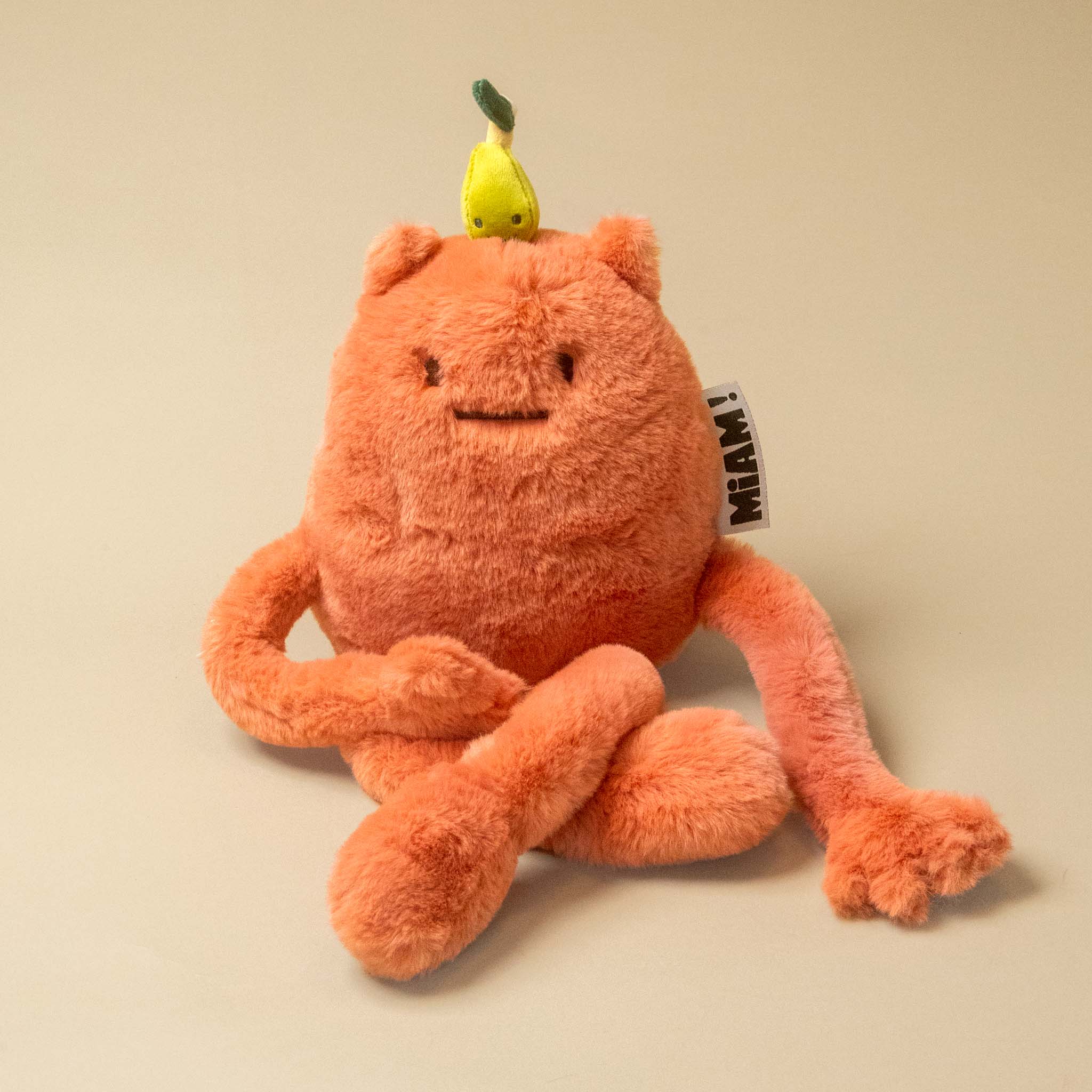 An orange plush britney-pear-petit toy with a small yellow pear on its head, photographed on a plain beige background