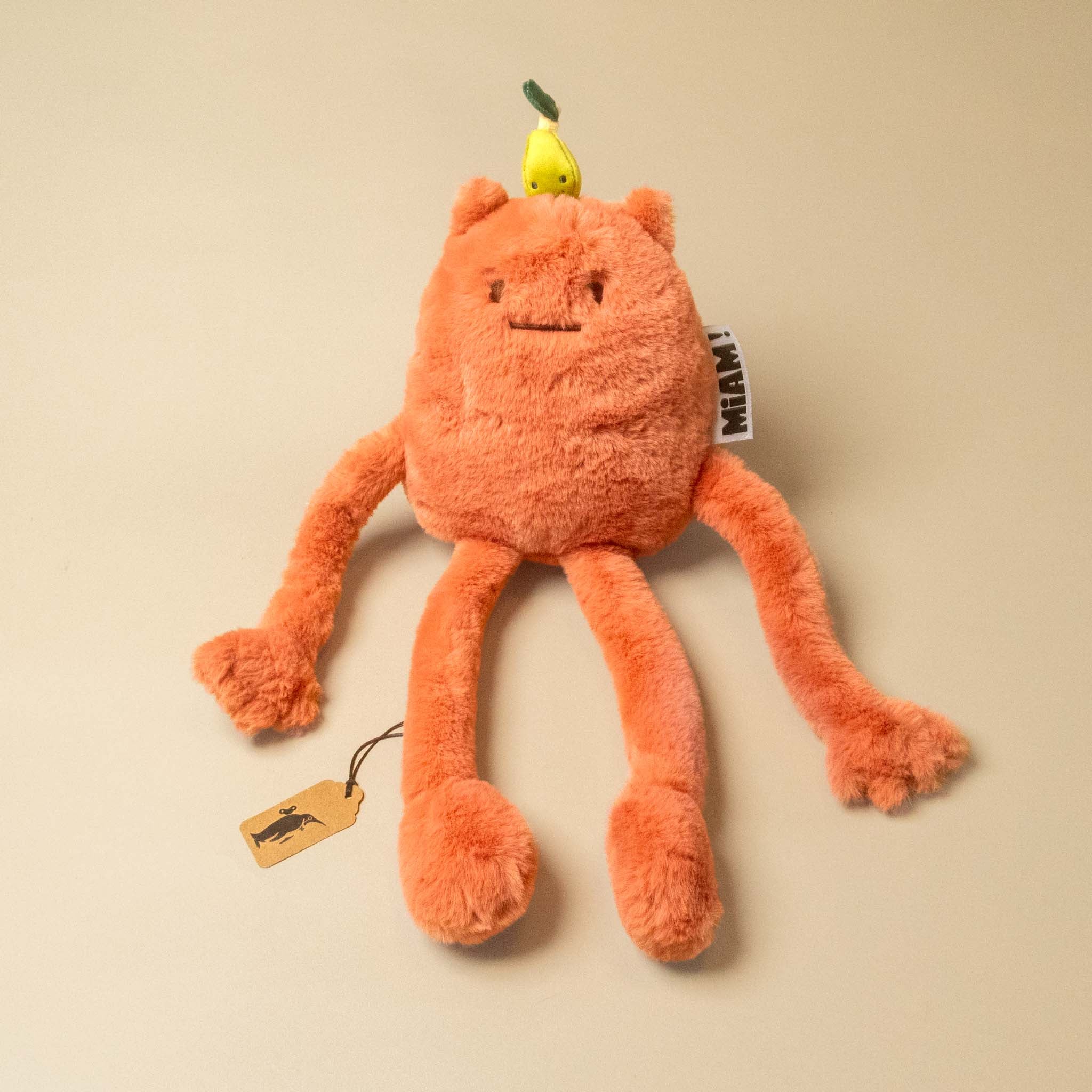 An orange plush britney-pear-petit toy with a small yellow pear on its head, photographed on a plain beige background