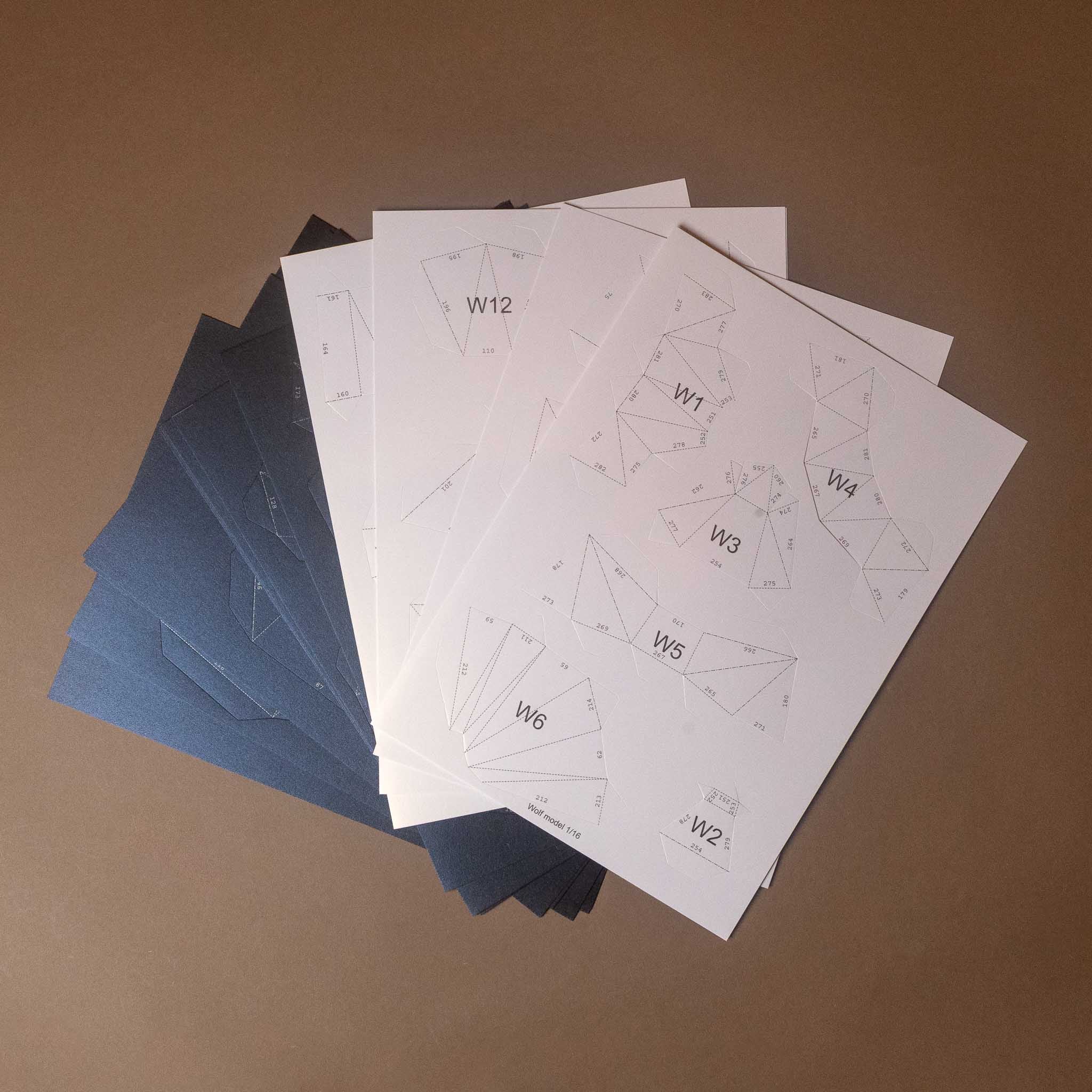 build-your-own-3-d-origami-wolf-lamp-model-kit-standing - stack of printed paper templates or pattern sheets fanned out on a plain brown background. The top sheet is white with faint geometric line drawings and labels such as W1 through W12, while sheets beneath are dark blue.