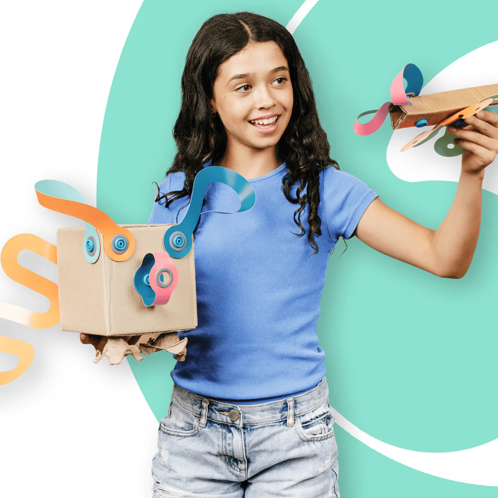 A smiling young girl with long dark hair stands against a teal and white graphic background, holding a cardboard box with colorful paper eyes and a tongue, and a small cardboard airplane in one hand.
