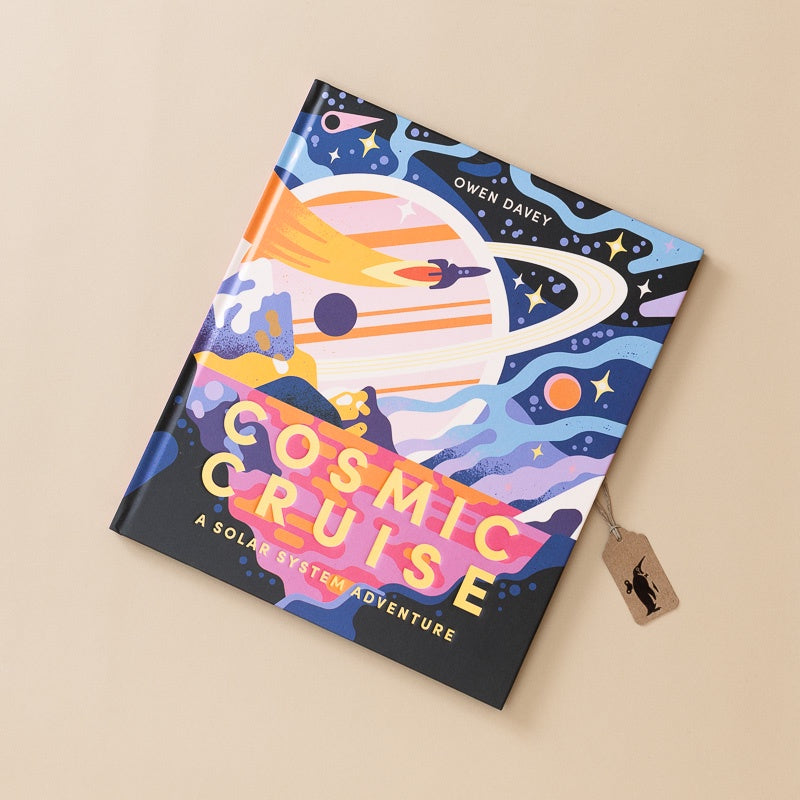 A hardcover book titled 'Cosmic Cruise: A Solar System Adventure' by Owen Davey, featuring a colorful retro-futuristic space illustration on the cover, photographed on a warm beige background with a small branded hangtag attached.