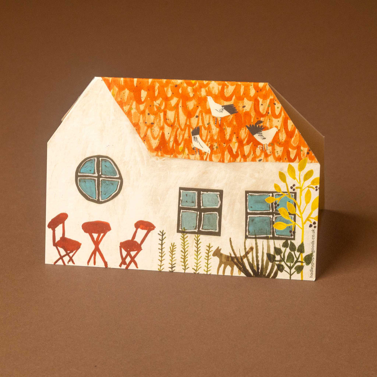 A small house-shaped decorative card or box with a hand-drawn illustration of a cottage, orange patterned roof, blue windows, red chairs, plants, and birds, photographed against a plain brown background.