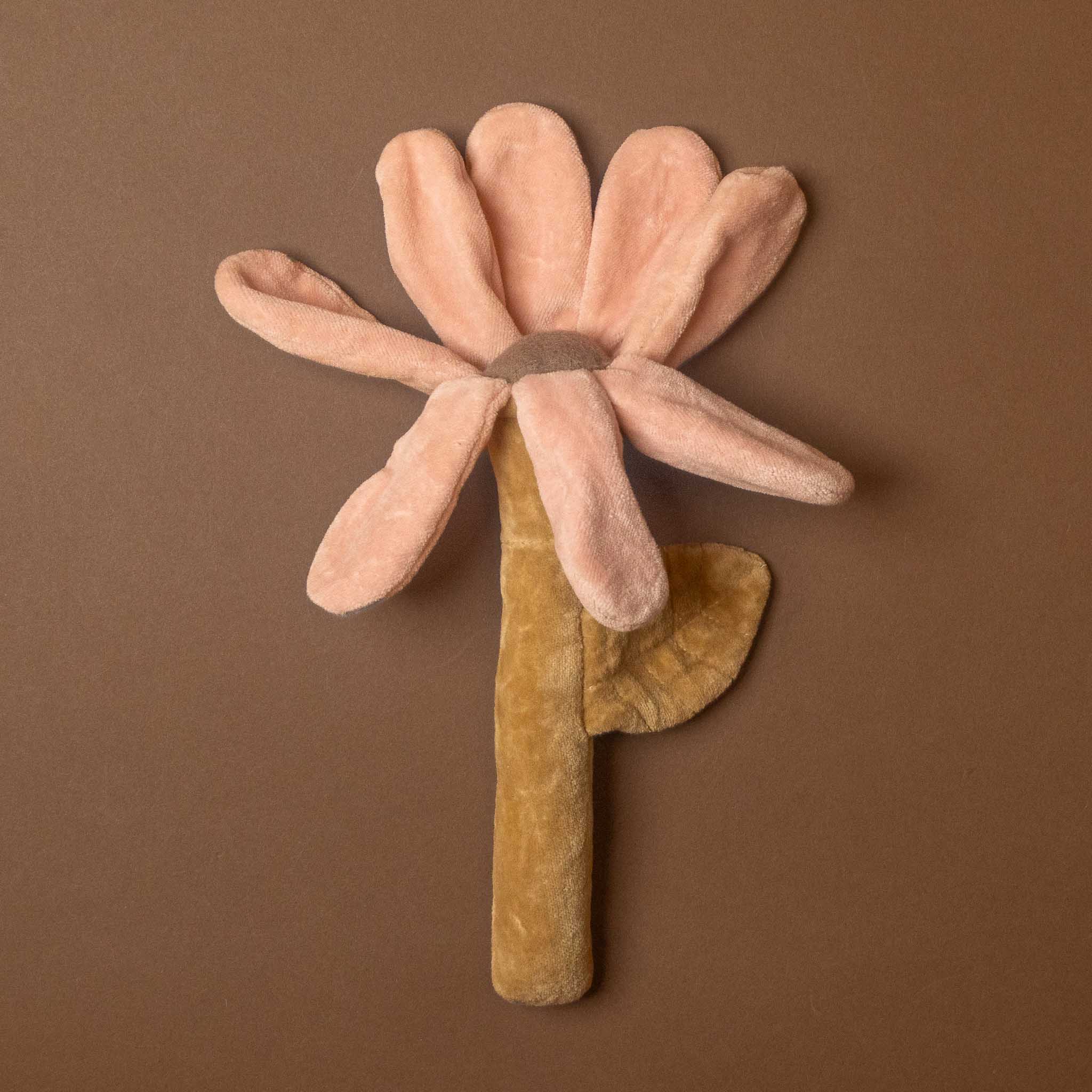 cotton-flower-rattle A plush flower-shaped product with soft blush pink petals, a tan center, and a mustard-yellow stem with a leaf, photographed against a solid brown background.