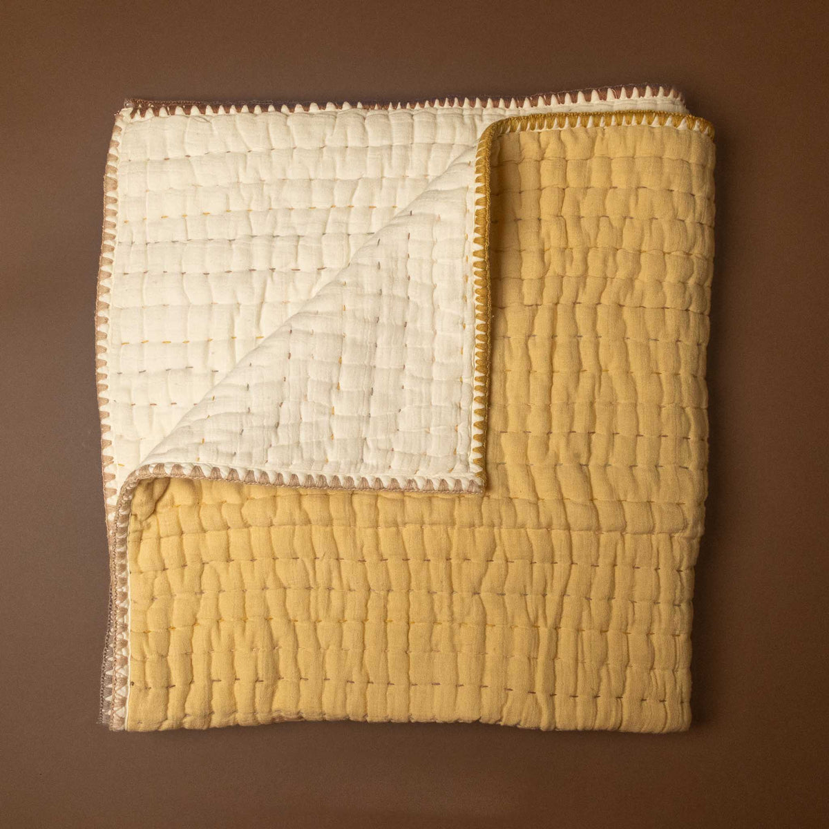 A folded quilted blanket or throw in two tones: a mustard yellow front side and an off-white reverse side, both with decorative zigzag stitching along the edges, displayed on a solid brown background.