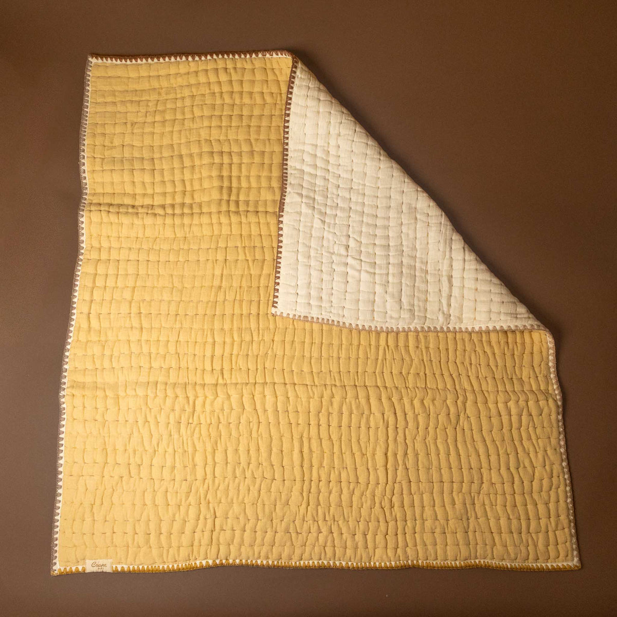 A quilted baby blanket with a mustard yellow front side and an off-white reverse side, laid flat on a solid brown background.