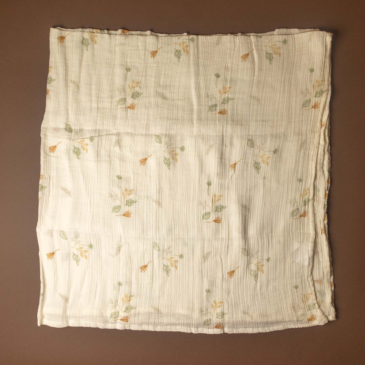 A square cream-colored muslin cloth with a delicate, small-scale floral pattern in muted green and orange, laid flat against a solid dark brown background.