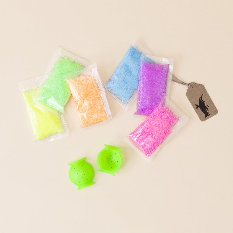 Six small clear plastic bags filled with brightly colored granular material in yellow, green, orange, blue, purple, and pink, arranged on a light beige background. Two small bright green round containers are placed in front, and a brown paper price tag hangs to the right.