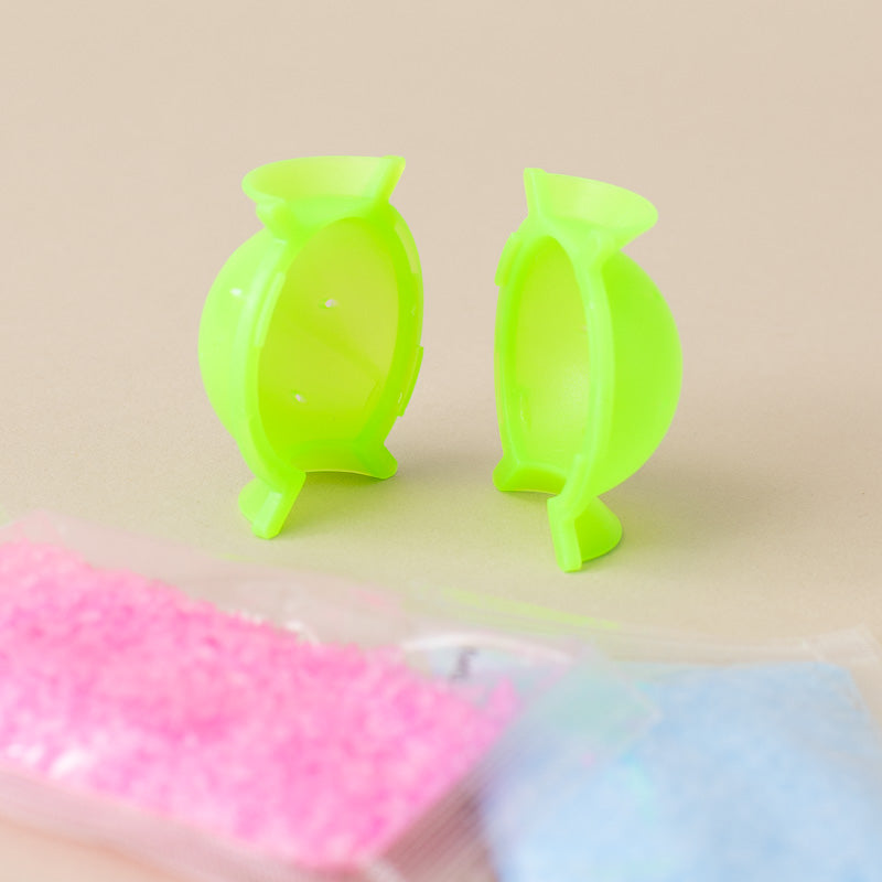 Two bright neon green plastic candy-shaped holders or molds standing upright on a beige background, with blurred pink and blue packets in the foreground.
