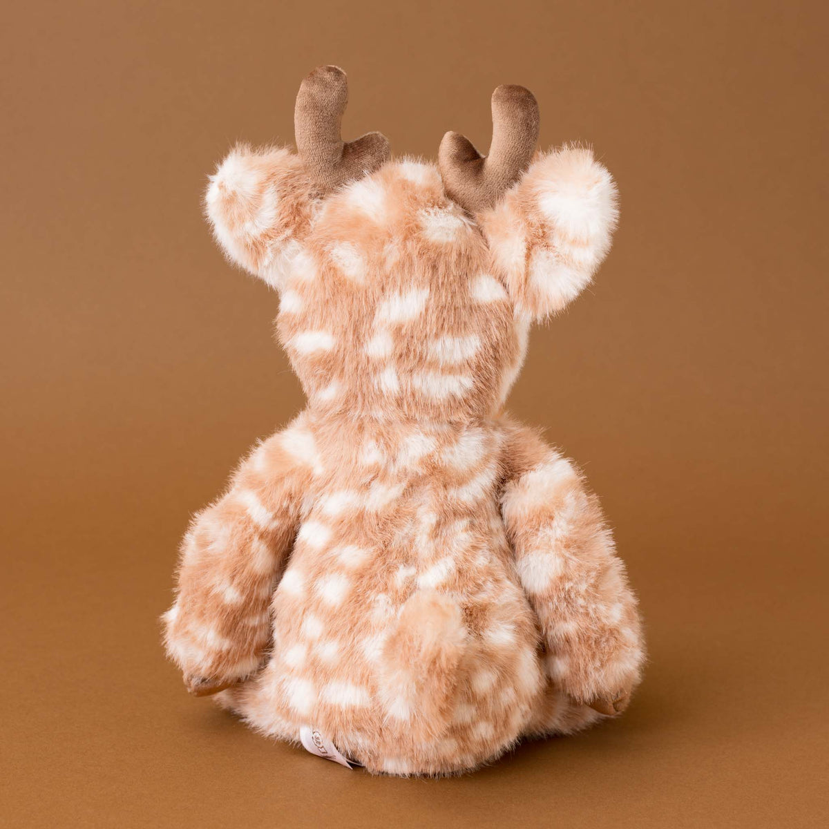 A plush stuffed animal shaped like a deer or antelope, shown from the back against a solid warm brown studio background. The toy has a soft tan and cream spotted fur pattern with brown antlers.