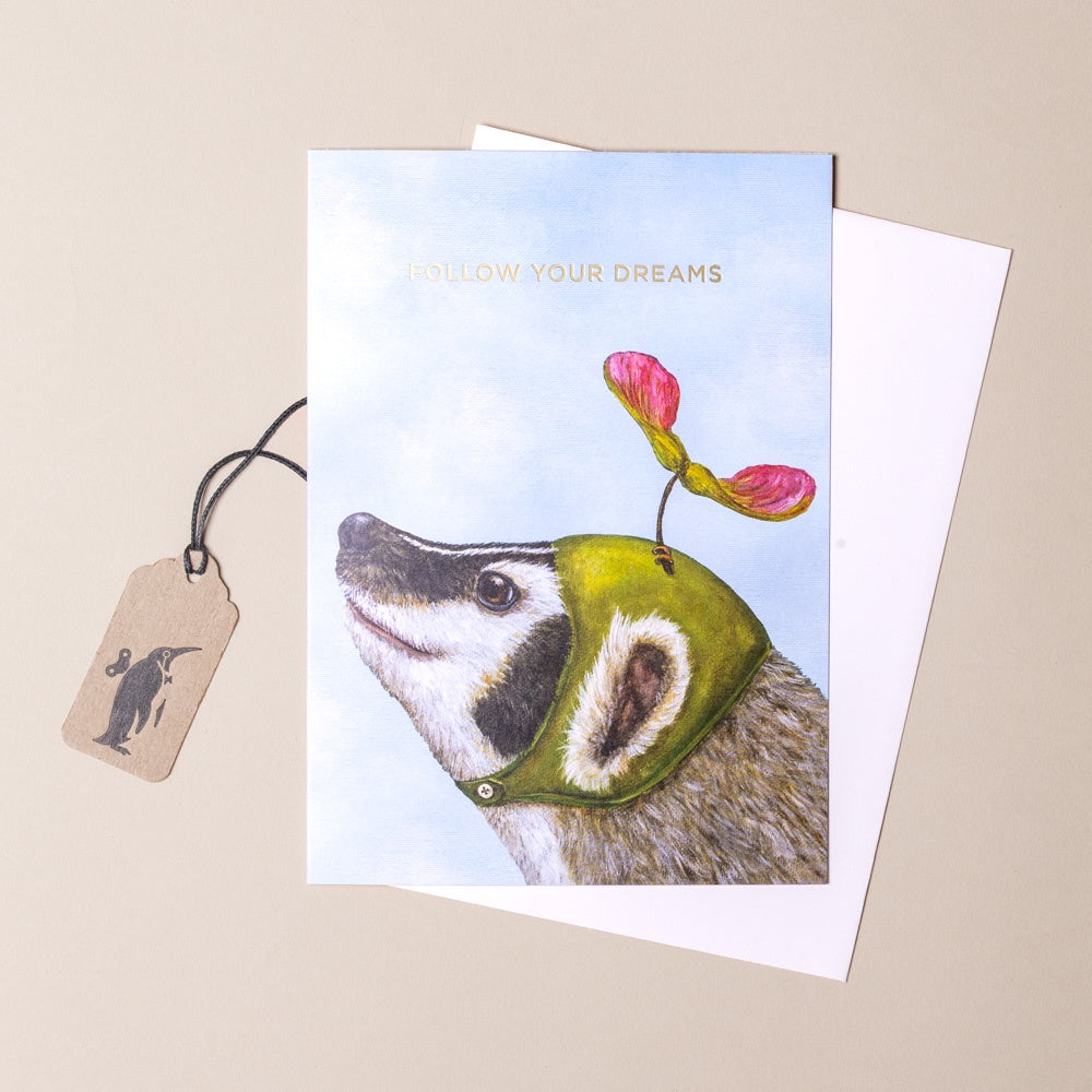 Greeting card with a badger wearing a green hat with flowers, accompanied by a tag on a beige background.
