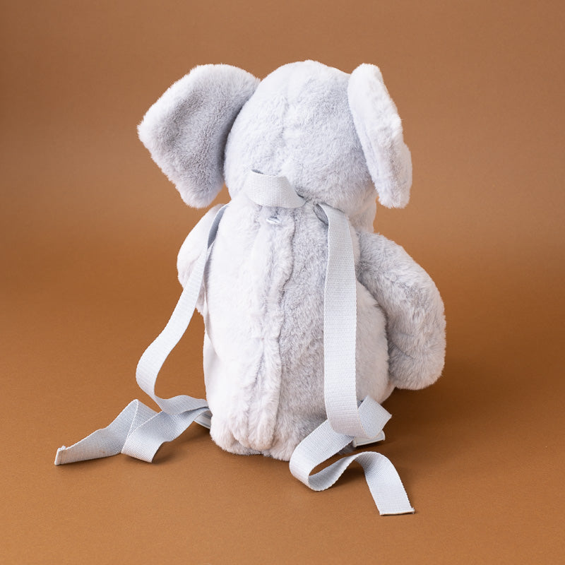 A plush elephant toy shown from the back, made of soft light gray fabric with long white ribbon tails tied around its neck, set against a solid warm brown background.