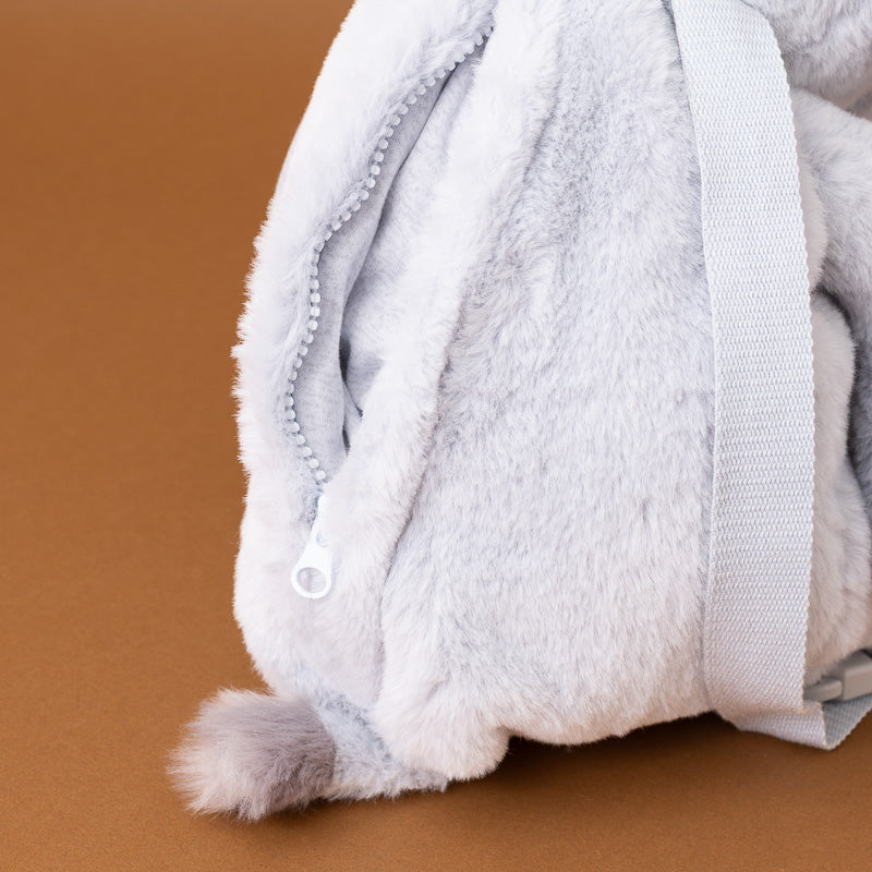 Close-up of a light gray plush fuzzy item with a zipper and a white webbed strap, photographed against a solid brown background.
