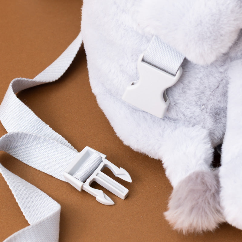 Close-up of a plush white stuffed animal or toy with soft faux-fur texture and white plastic buckle straps, laid on a brown surface.