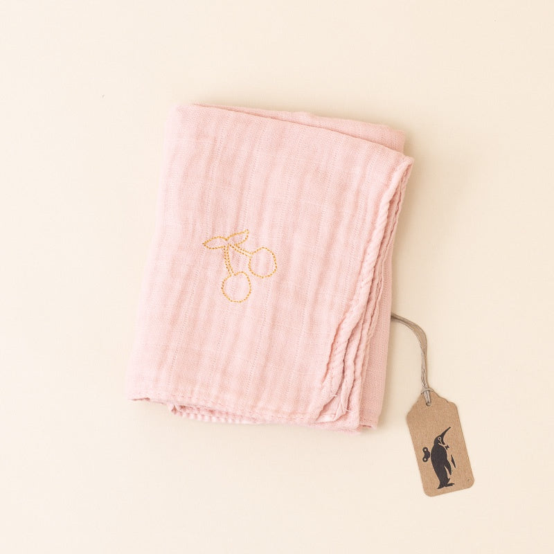 A folded light pink muslin cloth with a gold embroidered cherry design, shown on a warm cream background with a small kraft paper tag attached.