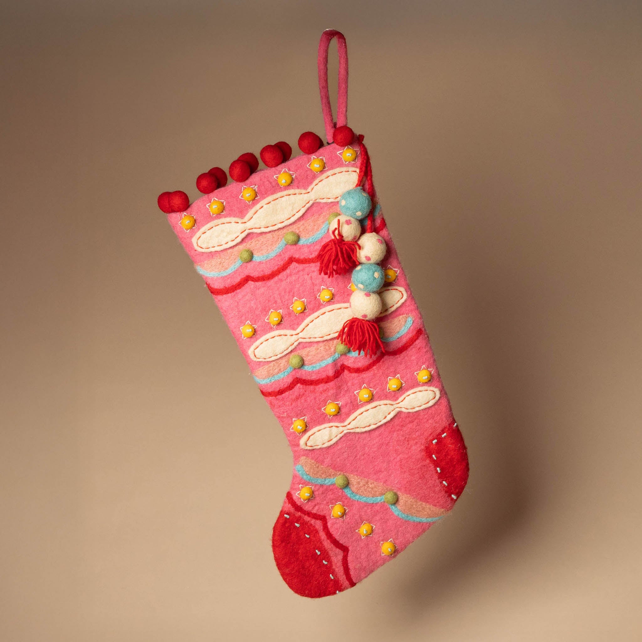 felted-evergreen-stocking-sugar-plum Decorative pink Christmas stocking with colorful tassels on a beige background