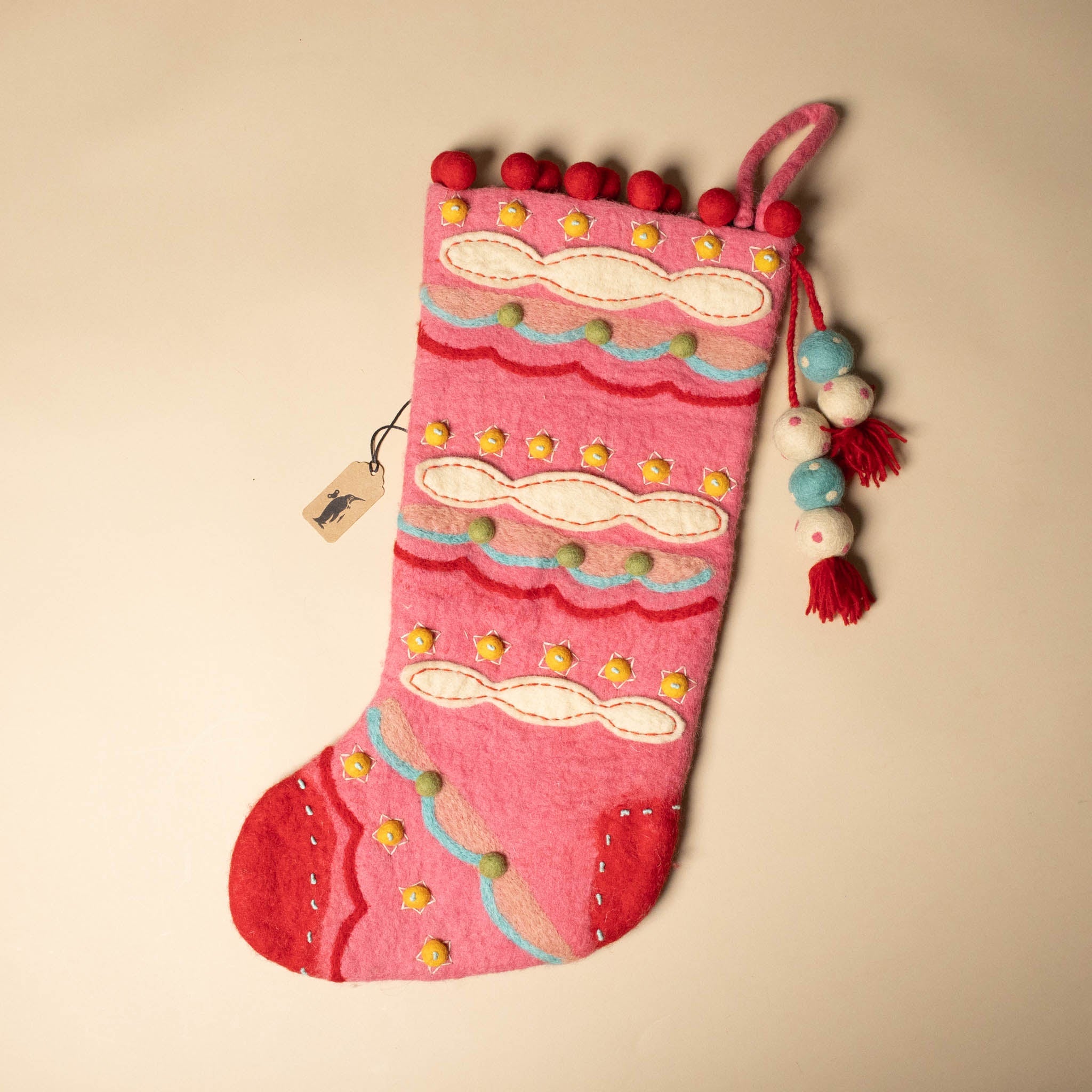felted-evergreen-stocking-sugar-plum Decorative pink Christmas stocking with colorful pom-poms on a beige background