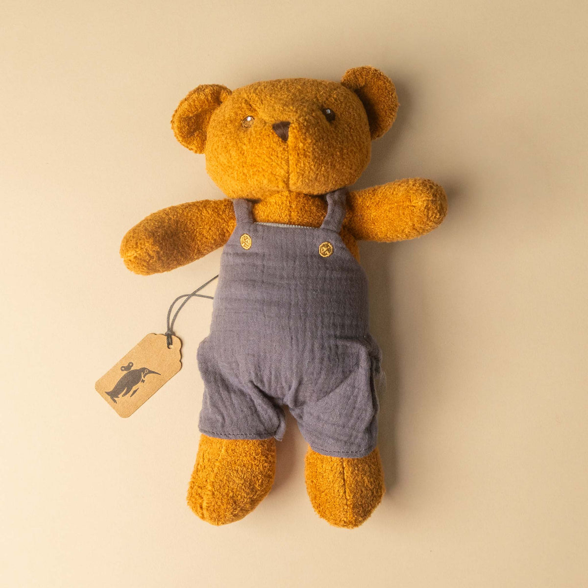 gabin-the-bear wearing blue overalls with a tag on a beige background