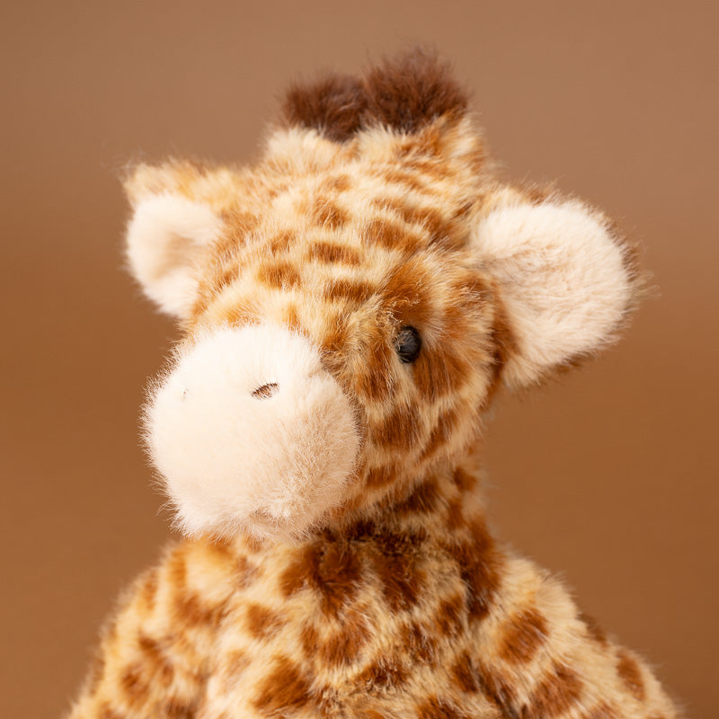 A close-up of a plush giraffe toy with brown and cream patches, a soft cream snout and ears, and a small dark eye, photographed against a warm brown studio background.