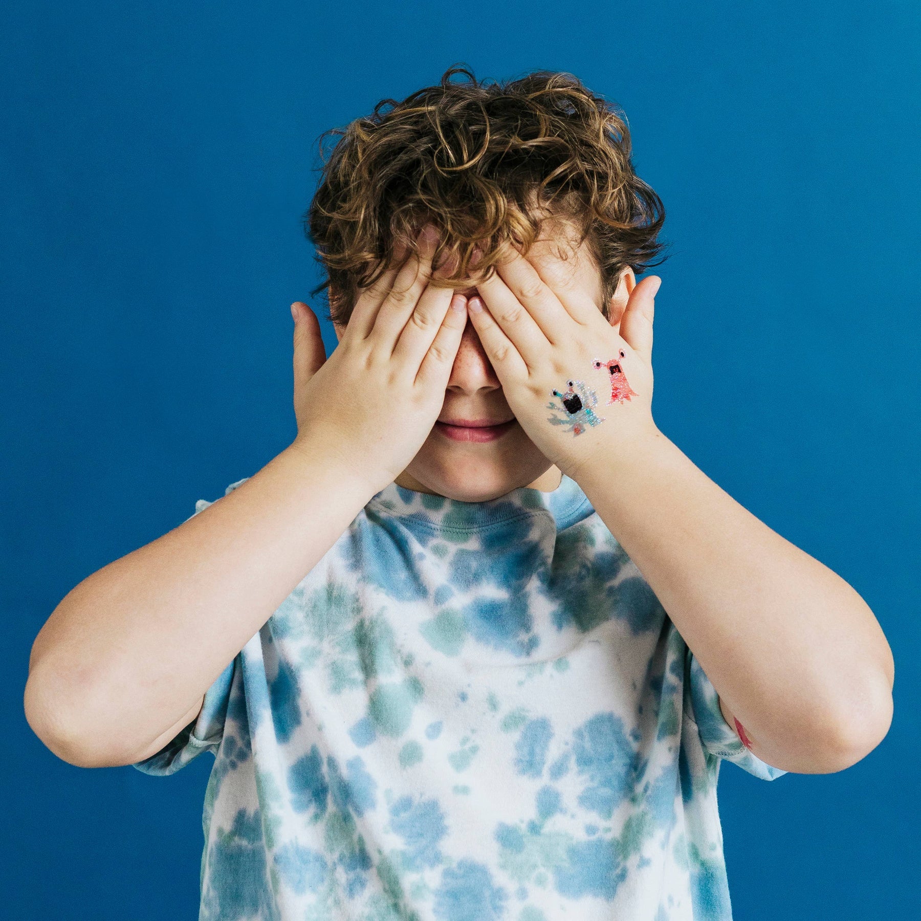 Child with hands over face glitter-monsters-temporary-tattoo-pairs against a blue background