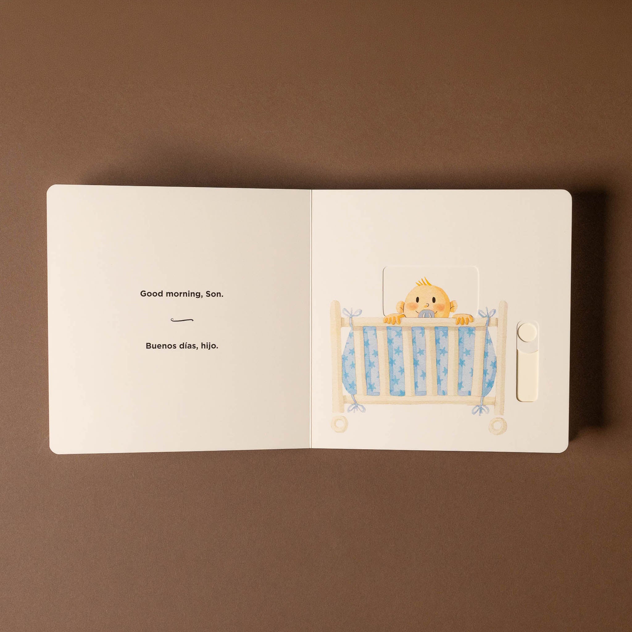 Open good-morning-buenos-dias-board-book with a illustration of a baby in a crib on a brown background