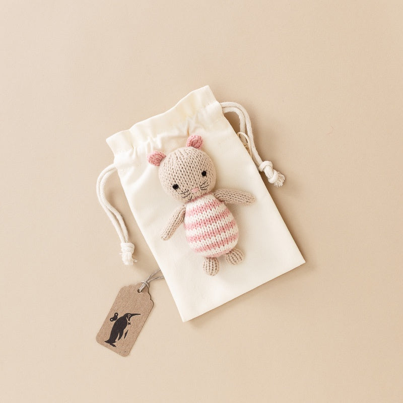 A small hand-knit-pocket-mouse with pink and white stripes and pink ears, resting on a cream drawstring pouch with a hanging kraft tag, photographed on a warm beige background.