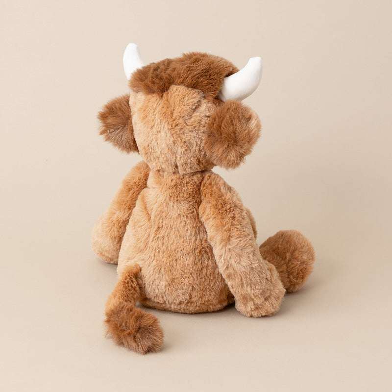 A plush toy bull seen from the back, featuring fluffy light brown fur, darker brown hair on its horns, and white horns, set against a solid beige background.