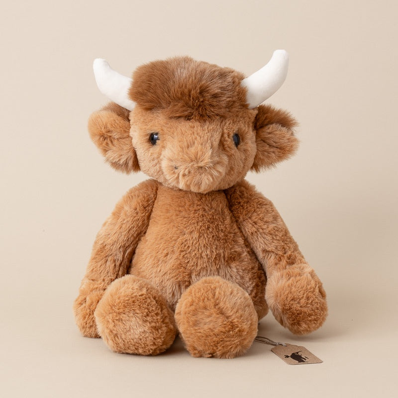 A plush Scottish Highland cow toy in warm brown faux fur with white horns, sitting against a solid beige background.