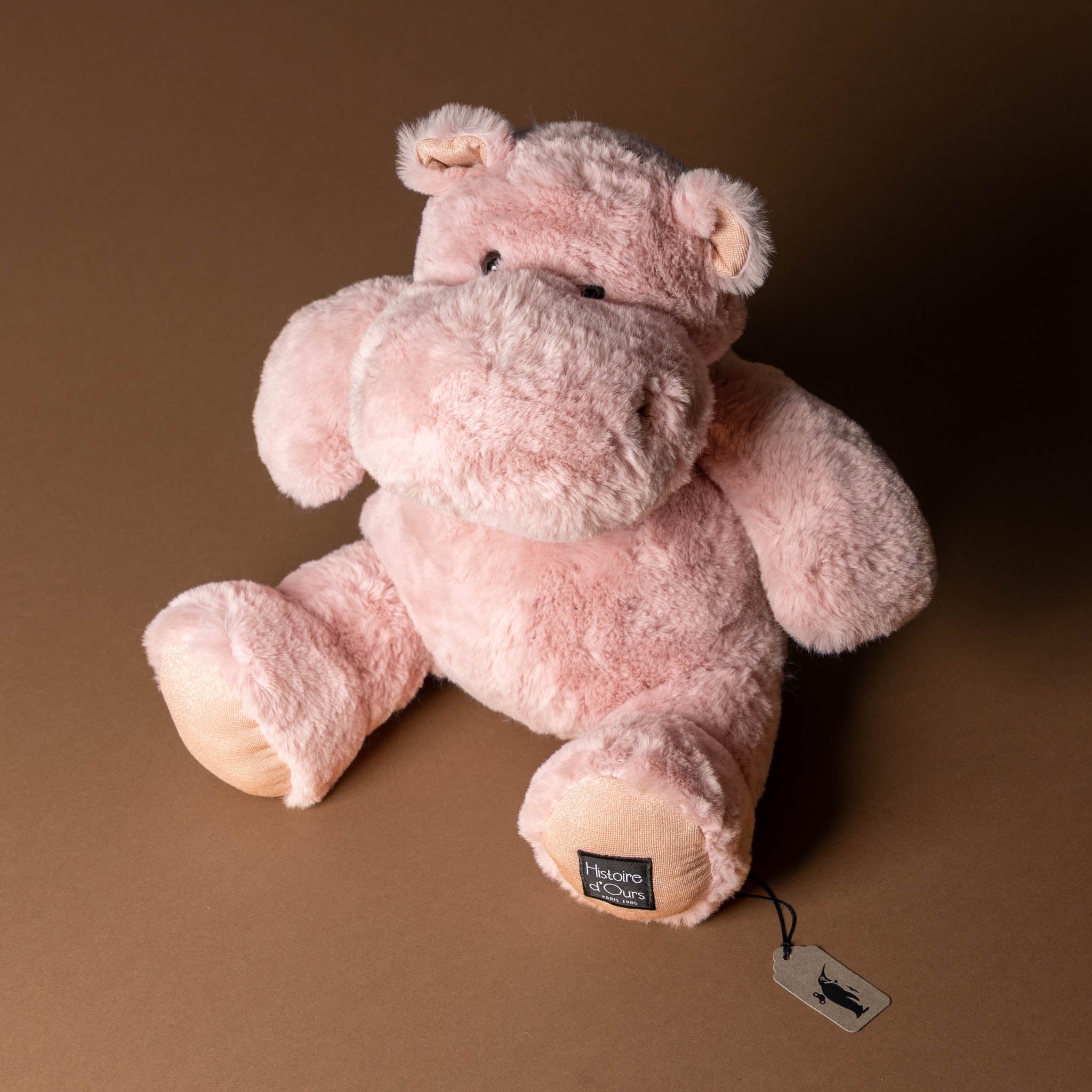 hip-hippo-blush-medium Pink plush hippo toy with a tag on a brown background