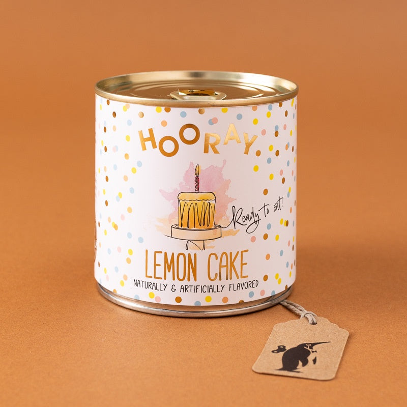 A cylindrical tin of 'Hooray Lemon Cake' ready-to-eat treats on a warm brown background, with a small paper hangtag attached.