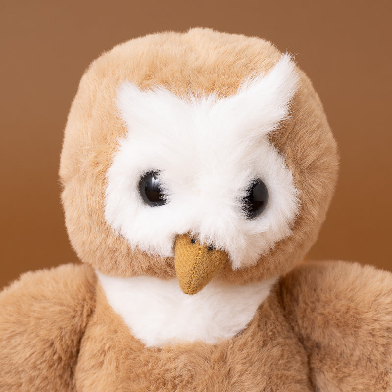 A close-up of a plush owl toy with fluffy tan-brown fur, a white face and chest, large black eyes, and a small tan beak, set against a solid warm brown background.
