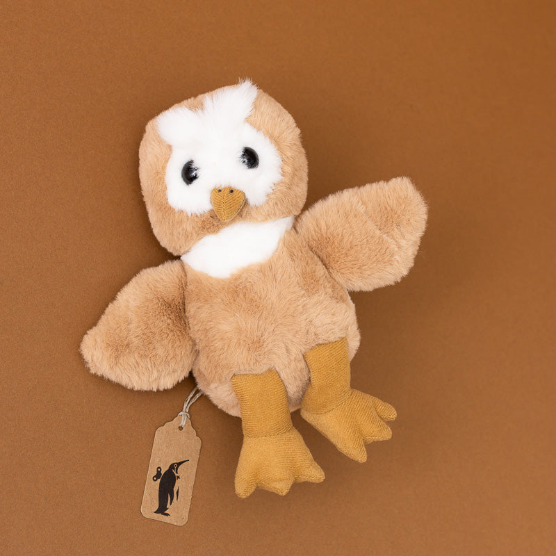 A small plush toy bird in tan and white with mustard-yellow feet and beak, photographed against a solid brown background. A small paper tag hangs from the side with a black bird silhouette logo.