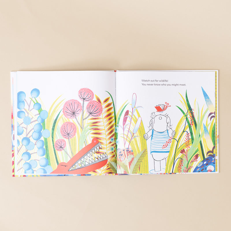 An open children's picture book laid flat on a warm beige background. The illustrated pages show colorful wildflowers, a red alligator, and a small character in tall grass with a butterfly.
