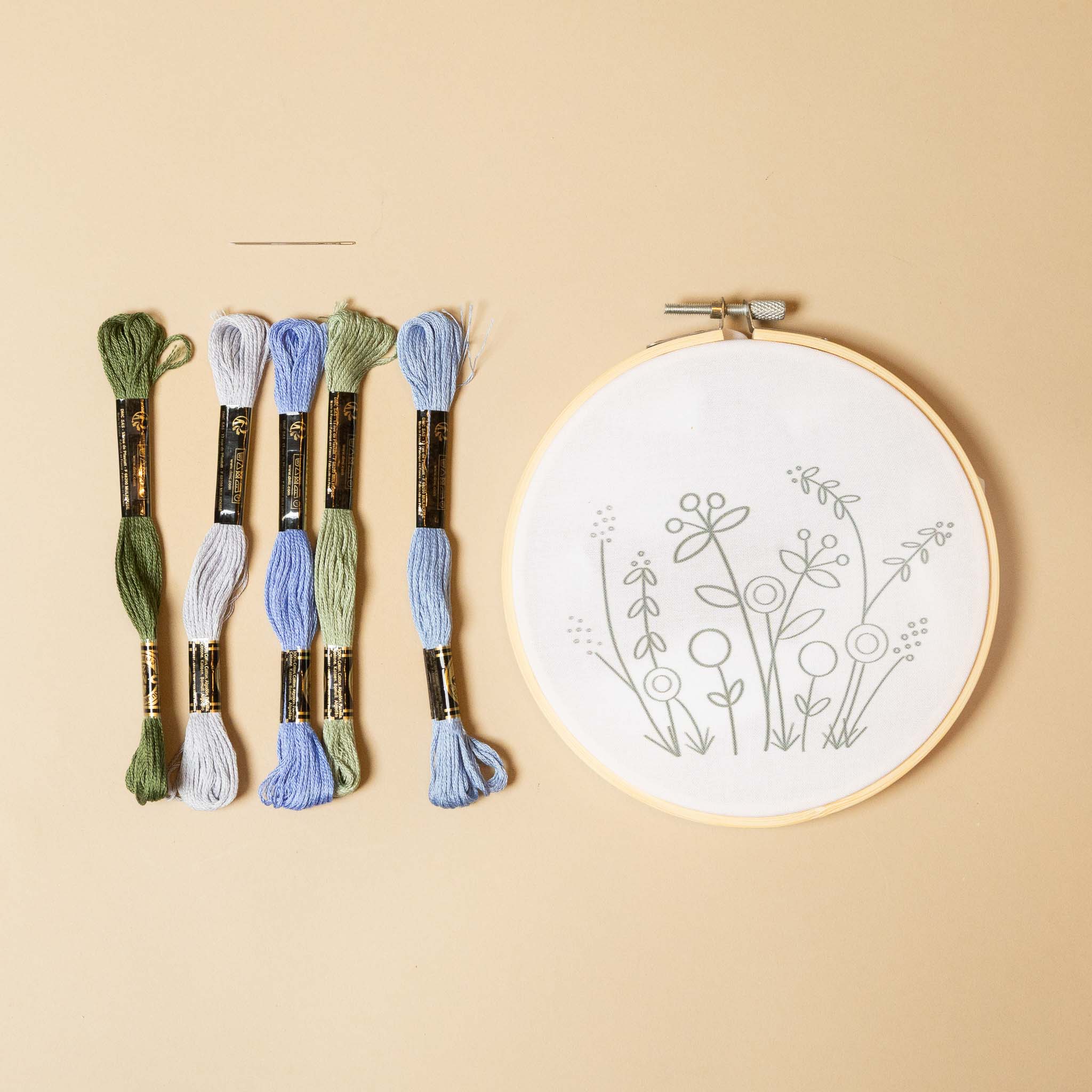 jardin-embroidery-kit-blues Embroidery hoop with floral pattern and set of colored threads on a beige background