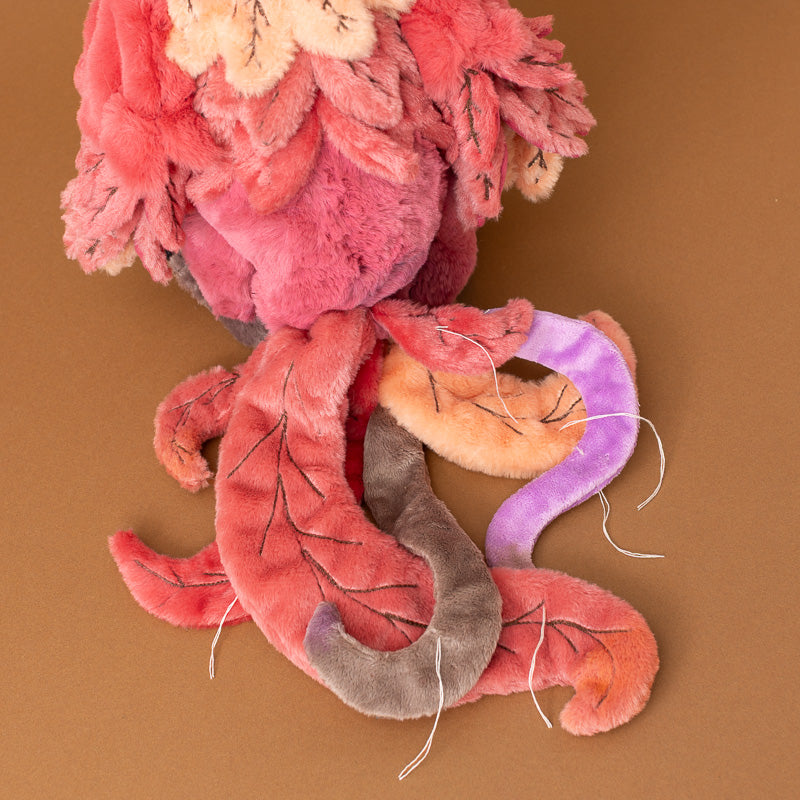 A plush jinzi-the-rising-phoenix toy with layered coral-pink and magenta fur, cream flower accents on its head, and four long tentacles in coral, orange, purple, and gray, photographed on a solid brown background.